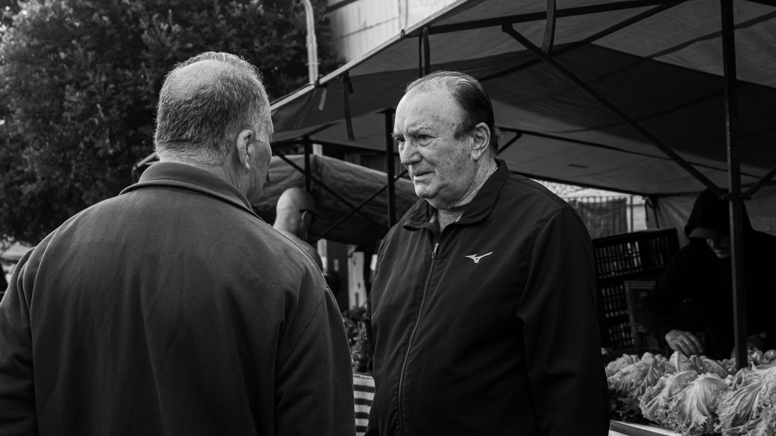 Duas pessoas conversam diante de uma banca coberta na feira.