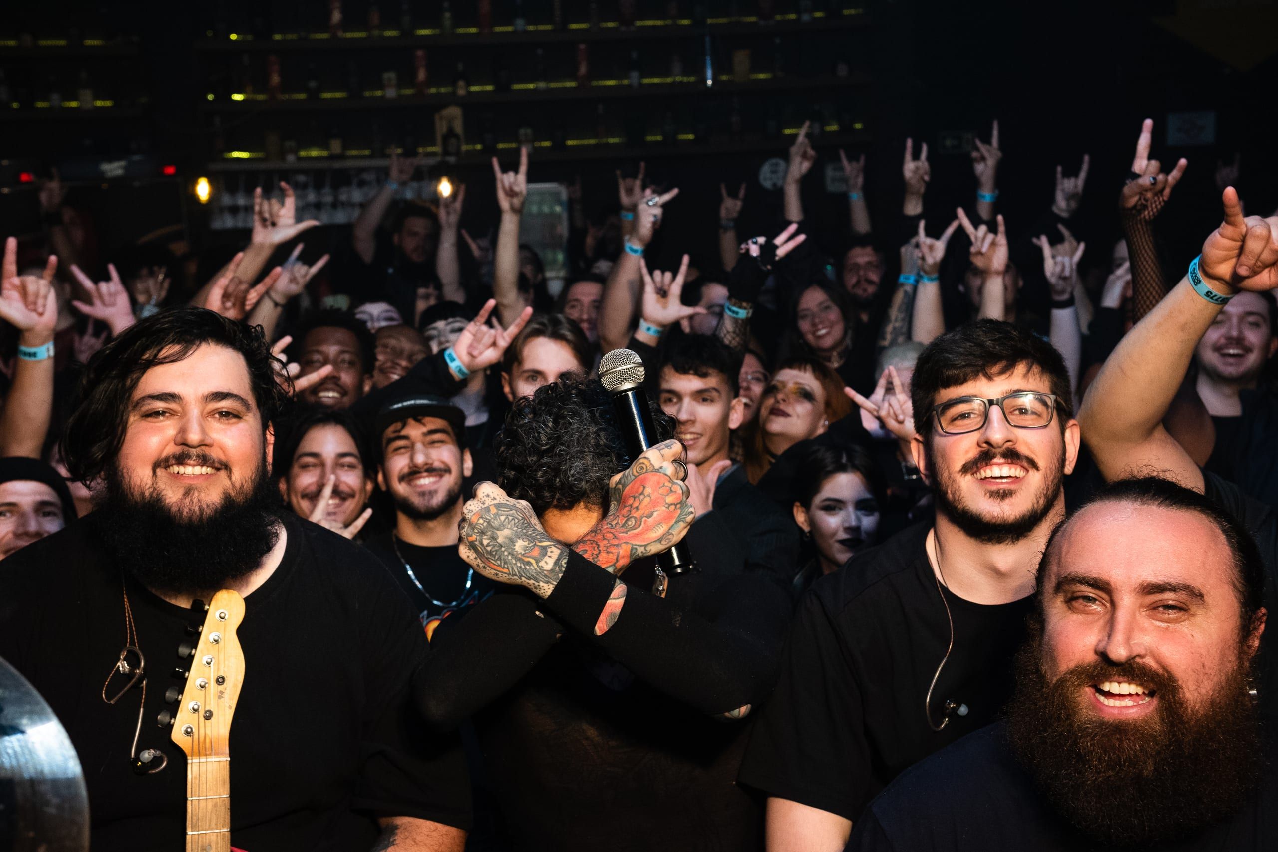 Grupo posa diante de um público com braços erguidos ao fundo.