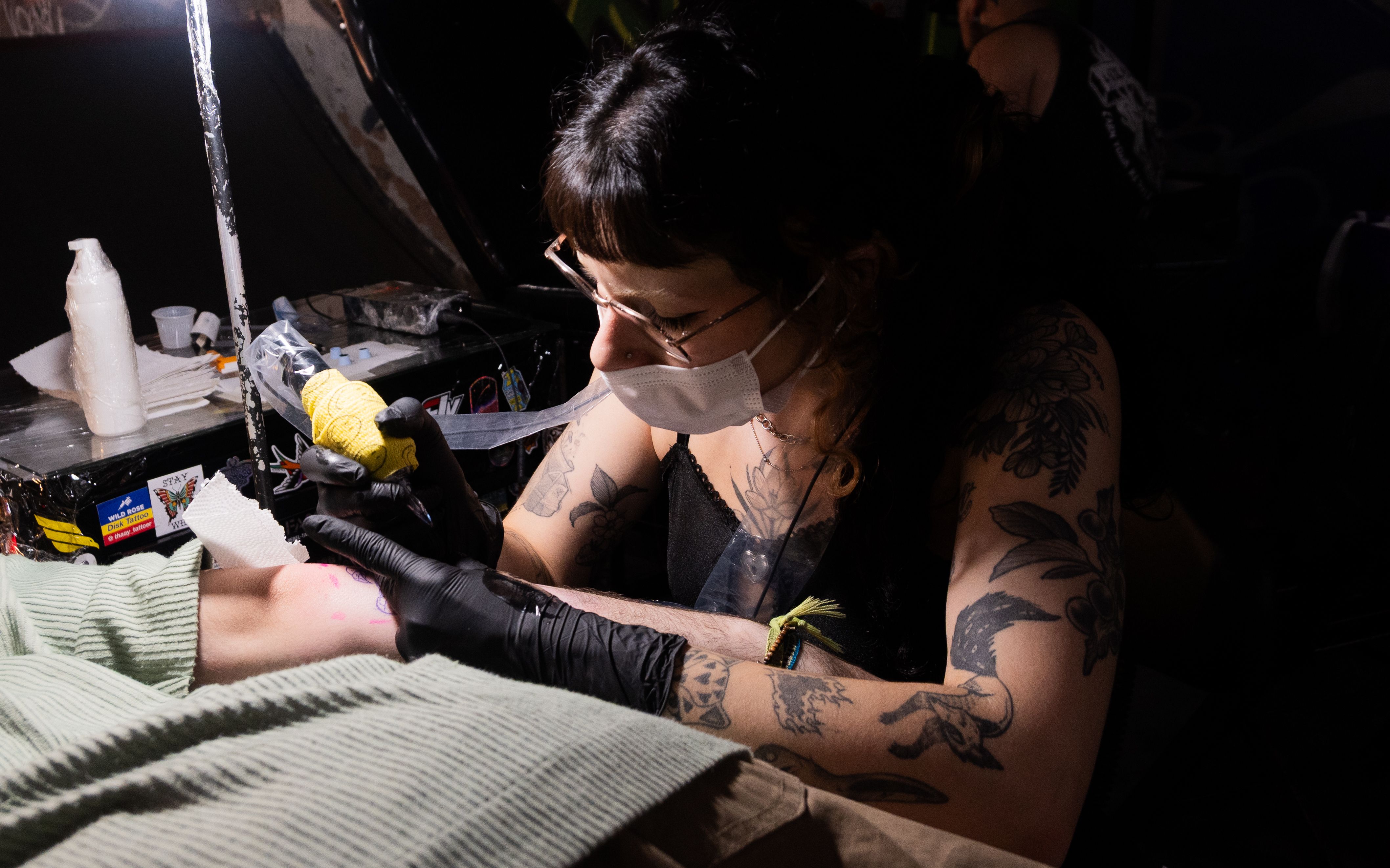 Pessoa tatuando o braço de outra pessoa em uma mesa de atendimento.