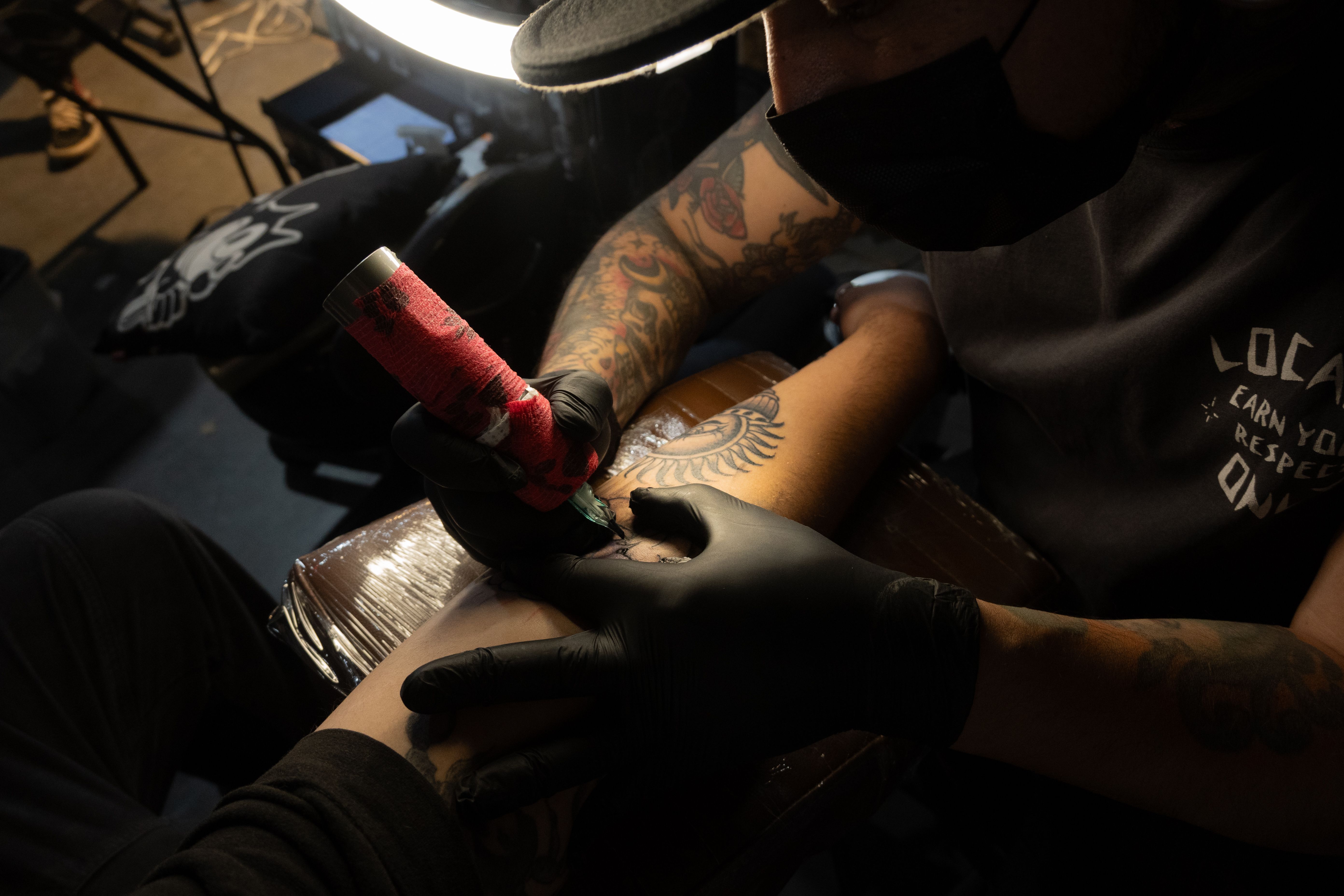 Mãos enluvadas seguram uma máquina de tatuagem sobre o braço de uma pessoa.
